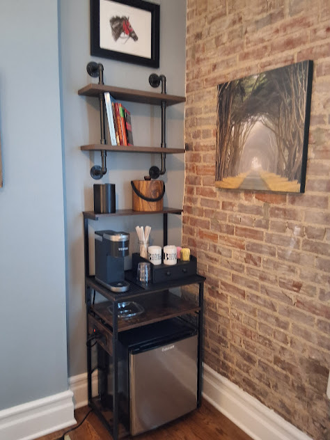 Corner with shelves, coffee station, and mini fridge next to wall of exposed brick.