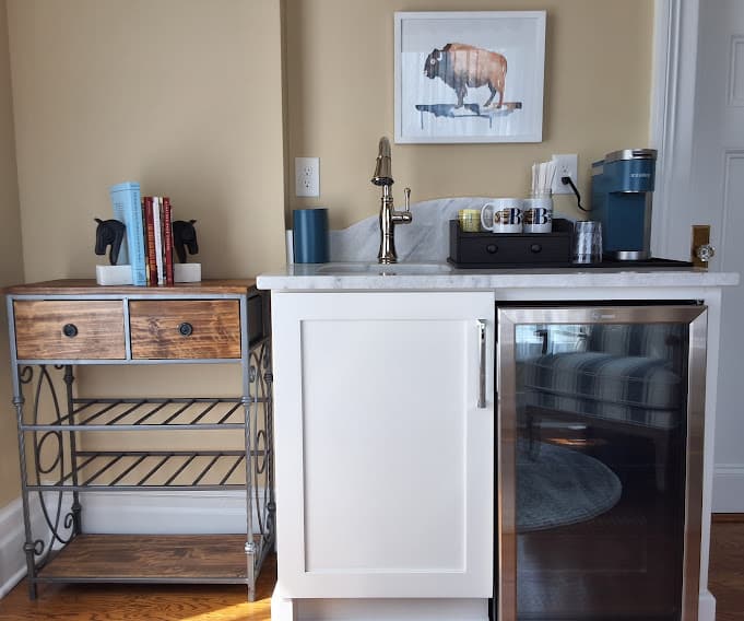Sink area in morning room with coffee maker, mugs, and mini-fridge next to book cart.