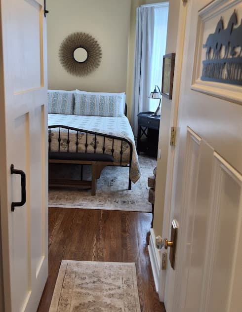 View of the Belmont Suite bedroom through the doorway revealing a metal king-size bed, hardwood floors, and area rugs.