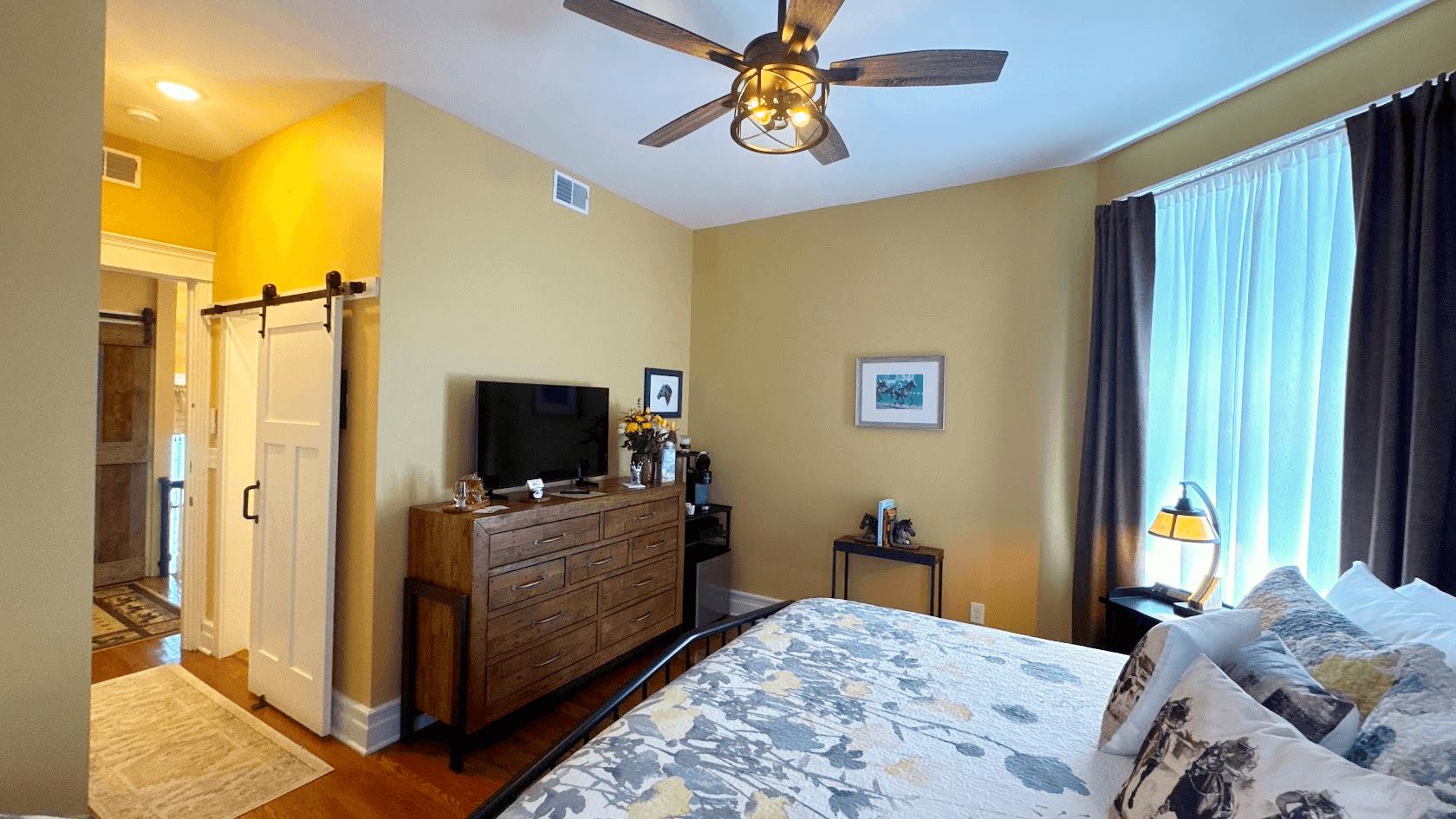 Tranquil bedroom with natural light, a king-size metal bed flanked by matching nightstands with lamps, hardwood floors, and area rug.