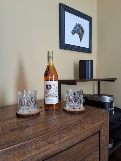Wooden dresser with two matching glasses and a bottle of bourbon next to coffee station.