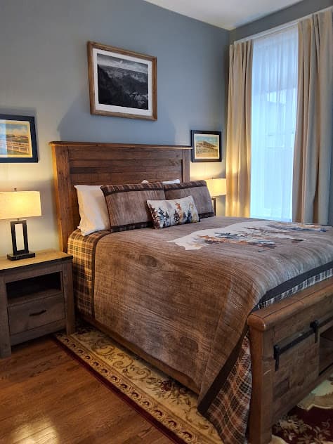 Cozy bedroom featuring a wooden bed with brown and plaid bedding, wooden nightstands with lamps, and framed artwork on gray walls.