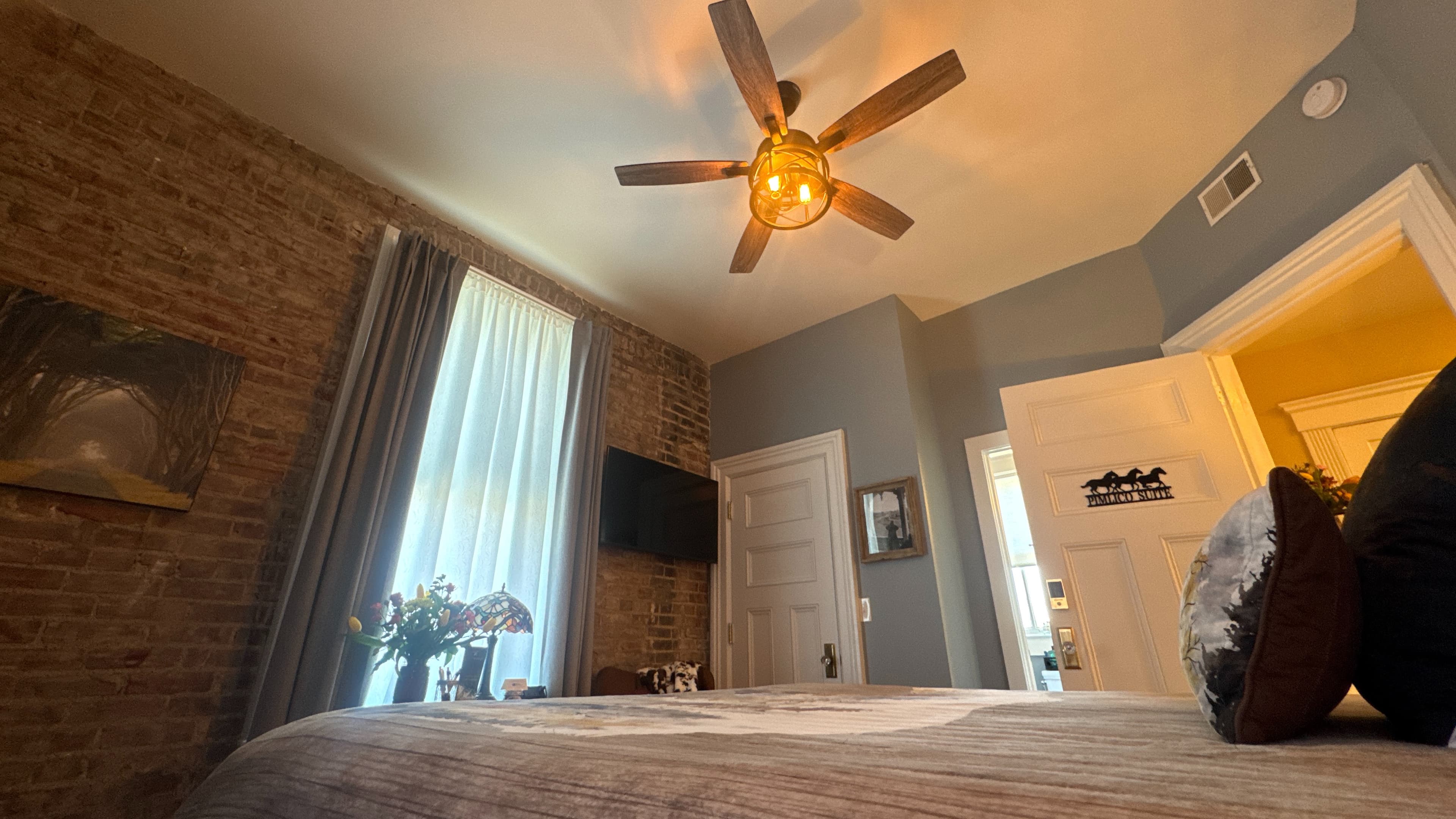 Bedroom featuring a ceiling fan, a bed with brown bedding, a window with curtains, a brick wall with a mounted TV, and a wooden floor.