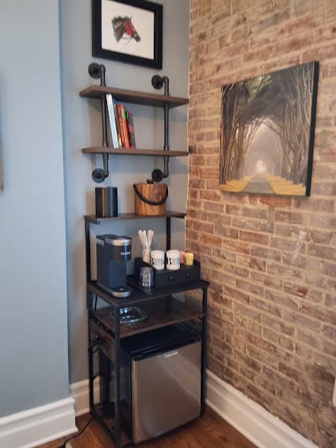Corner with shelves, coffee station, and mini fridge next to wall of exposed brick.
