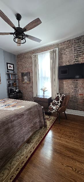 Bedroom featuring a ceiling fan, a bed with brown bedding, a window with curtains, a brick wall with a mounted TV, and a wooden floor.