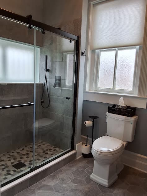 A bathroom featuring a walk-in shower with glass doors, a white toilet, a window with a blind, and gray hexagonal floor tiles.