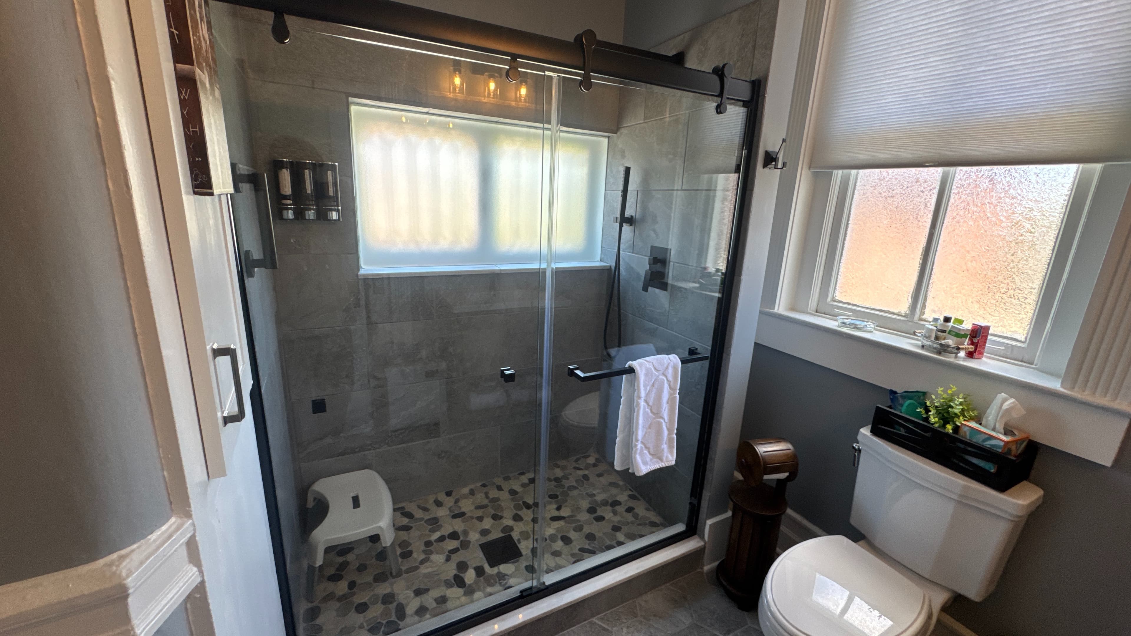 A bathroom featuring a walk-in shower with glass doors, a white toilet, a window with a blind, and gray hexagonal floor tiles.