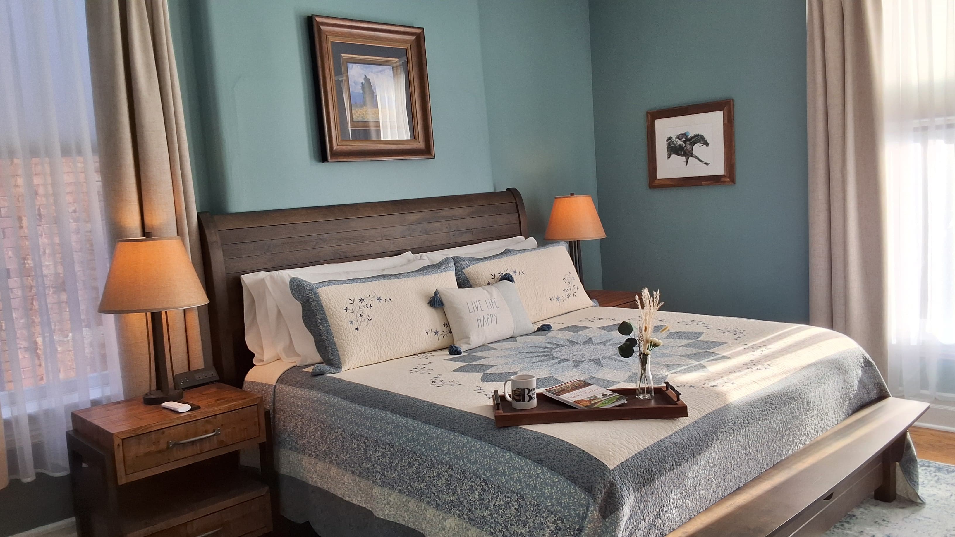 A tranquil bedroom decorated in blues and neutrals with a king-size bed topped with a quilt, hardwood floors with area rug, a ceiling fan, and a window letting in plenty of natural light.