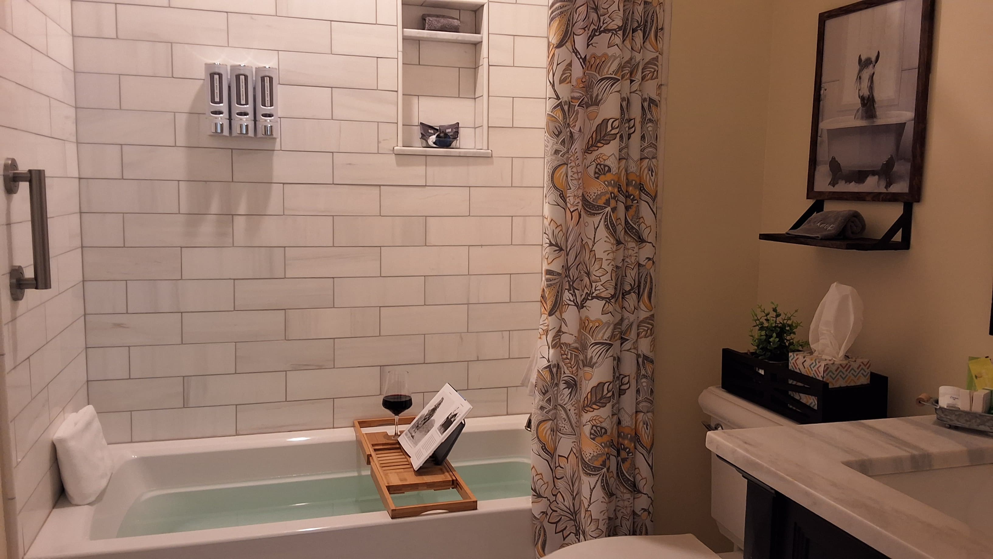 Cozy bathroom with a filled tub, bath tray, wine glass, book, and floral shower curtain