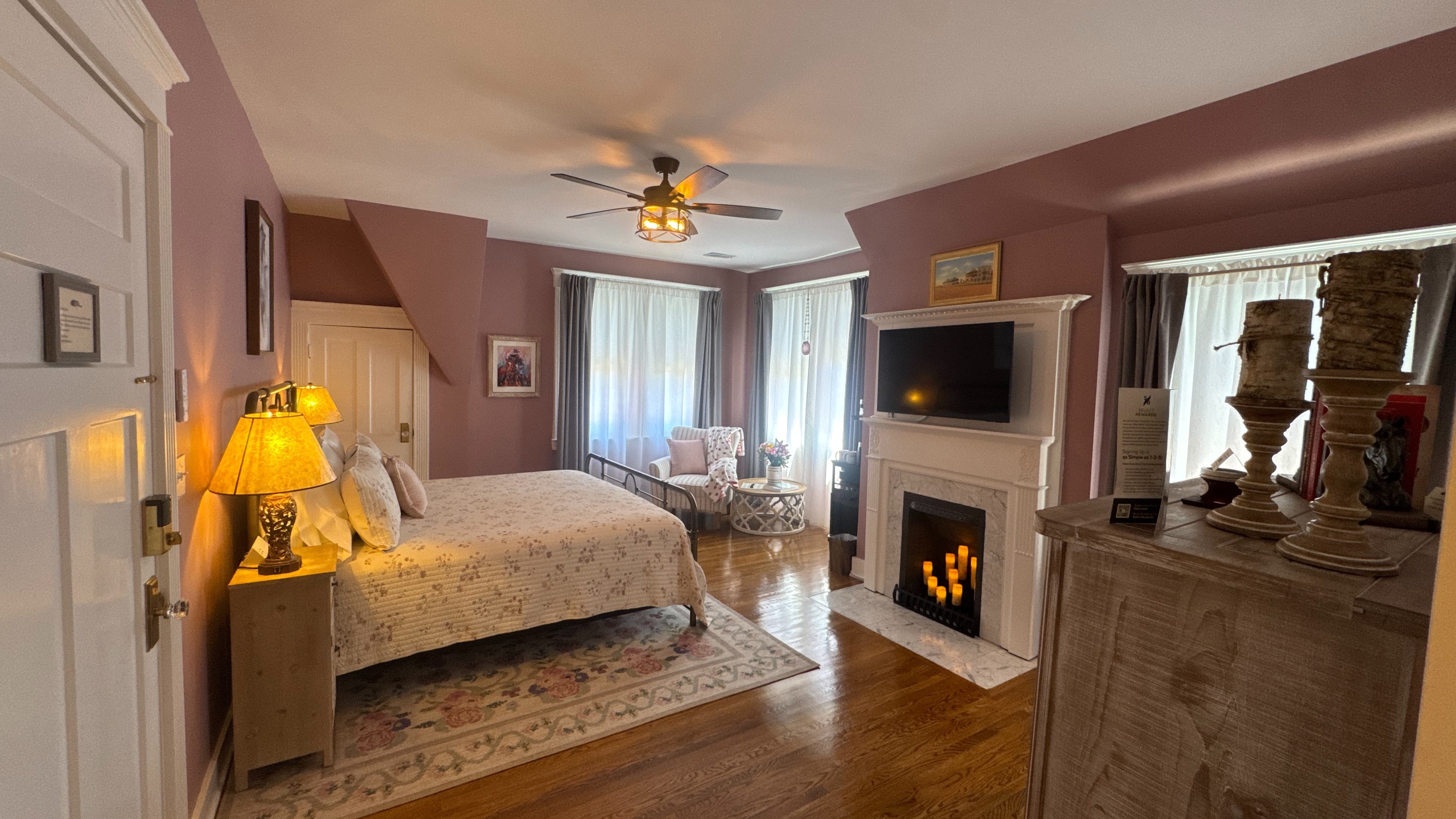 Charming bedroom with king-size bed, decorative fireplace, flat-screen TV, hardwood floors, and ceiling fan.