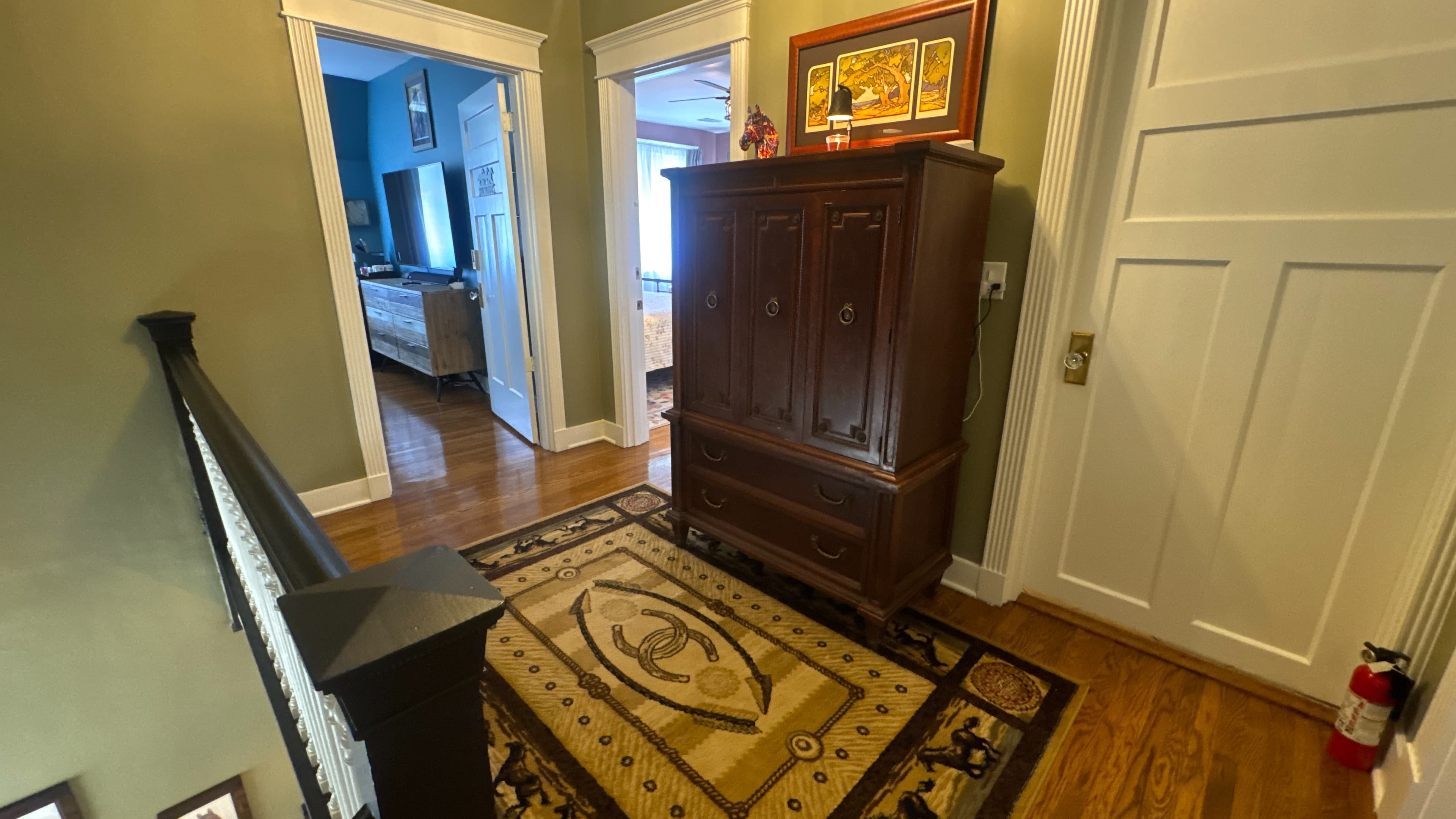 A wooden cabinet sits on a patterned rug in a well-lit hallway leading to adjacent rooms.