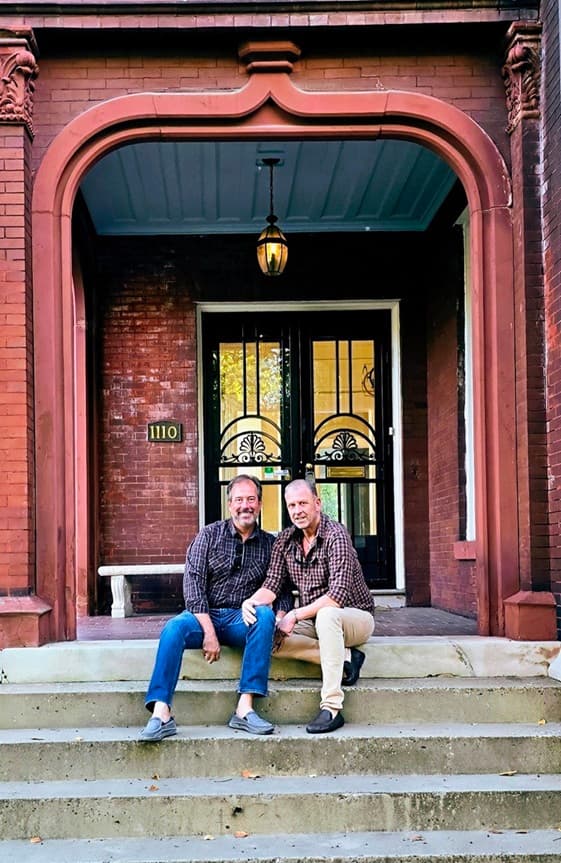 Two men sit on the steps of a building, smiling at the camera.