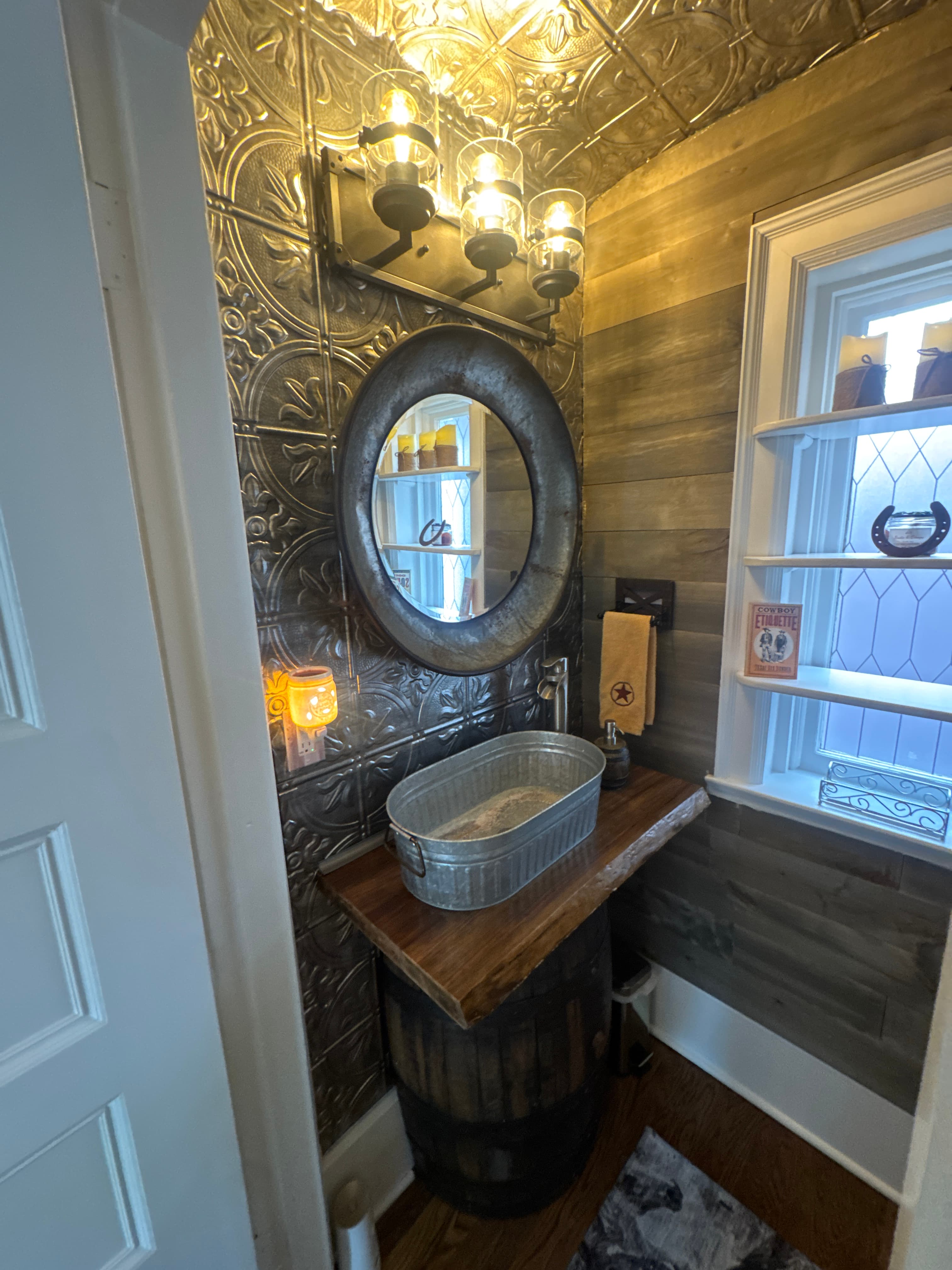 A rustic bathroom corner featuring a metal mirror, wooden shelf, and a basin atop a barrel.