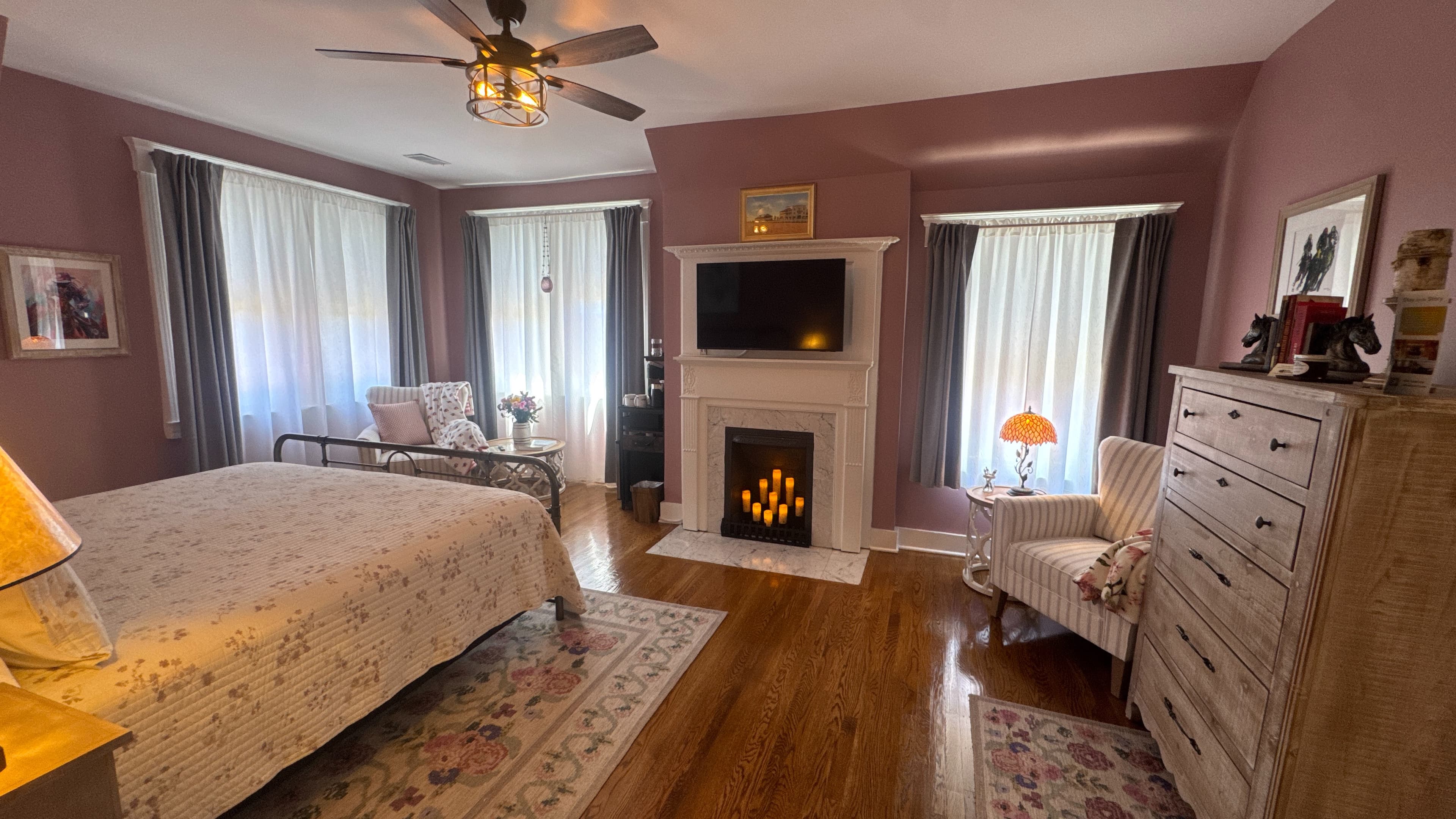 Charming bedroom with king-size bed, decorative fireplace, flat-screen TV, hardwood floors, and ceiling fan.