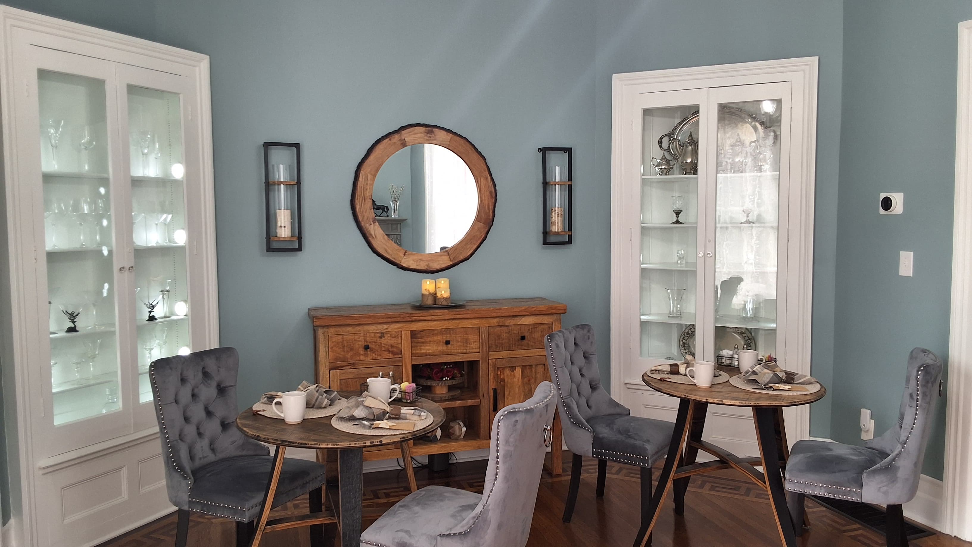 A cozy dining area features two round tables with gray chairs, a wooden sideboard, and decorative glassware.