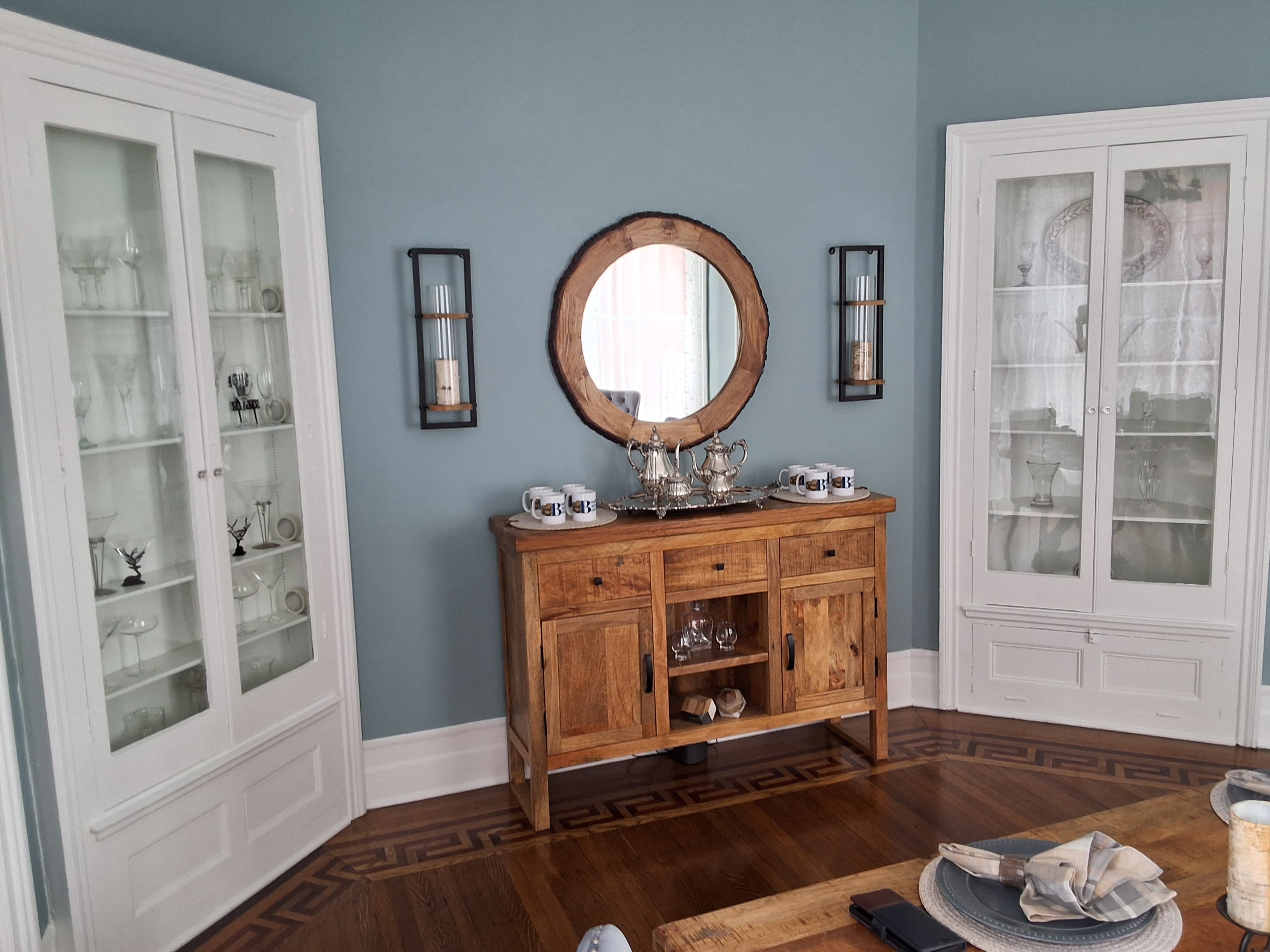 A cozy dining area featuring wooden furniture, glass display cabinets, and a round mirror against a light blue wall.
