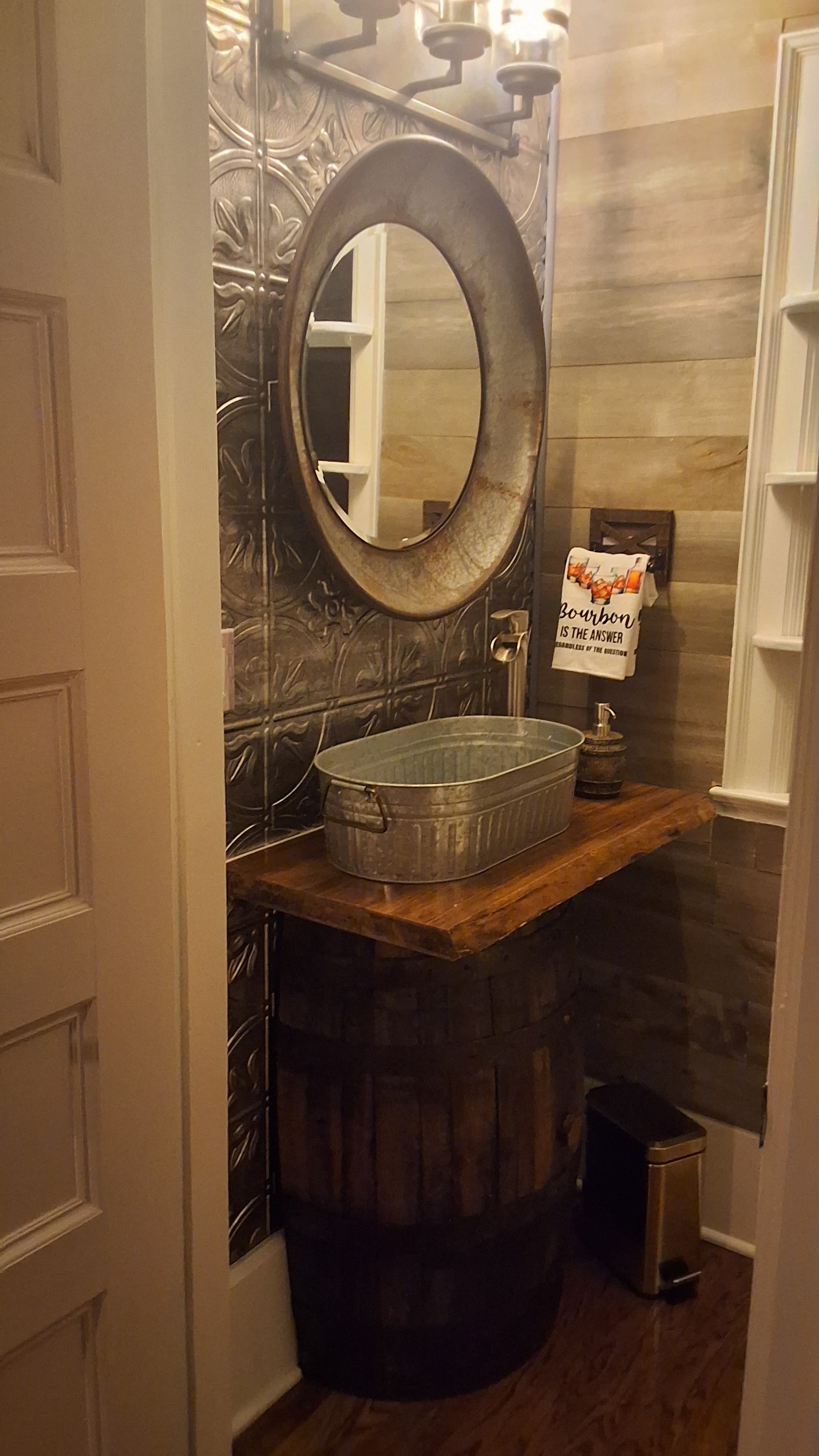 A rustic bathroom corner featuring a wooden table on a barrel, a galvanized sink, and a round mirror.