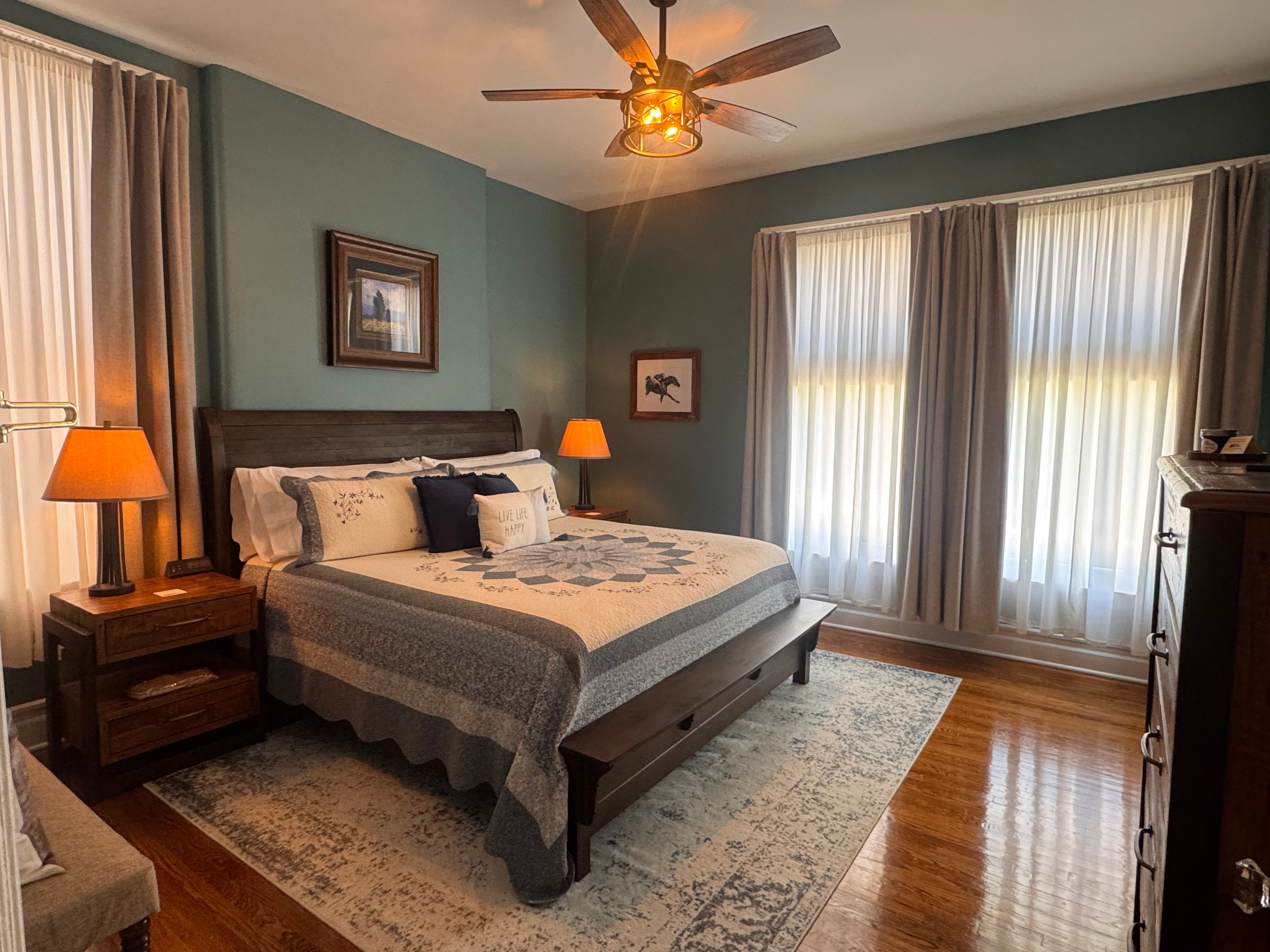 A tranquil bedroom decorated in blues and neutrals with a king-size bed topped with a quilt, hardwood floors with area rug, a ceiling fan, and a window letting in plenty of natural light.